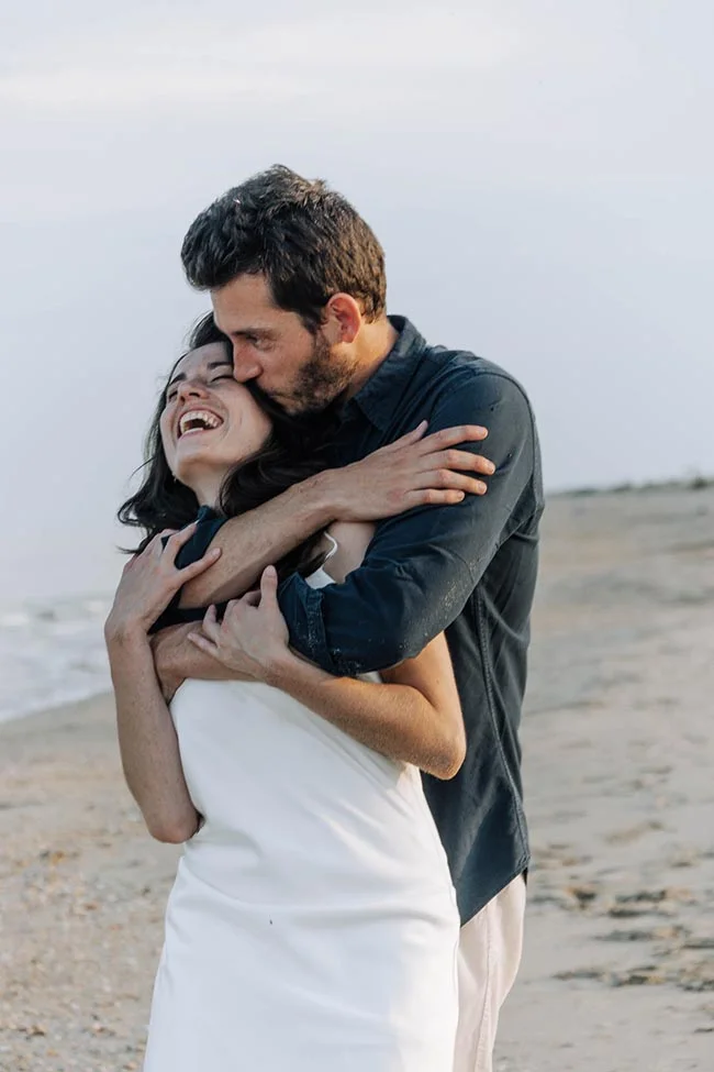 Un couple s'enlace avec complicité sur une plage, la mariée en robe blanche riant aux éclats dans les bras de son époux.