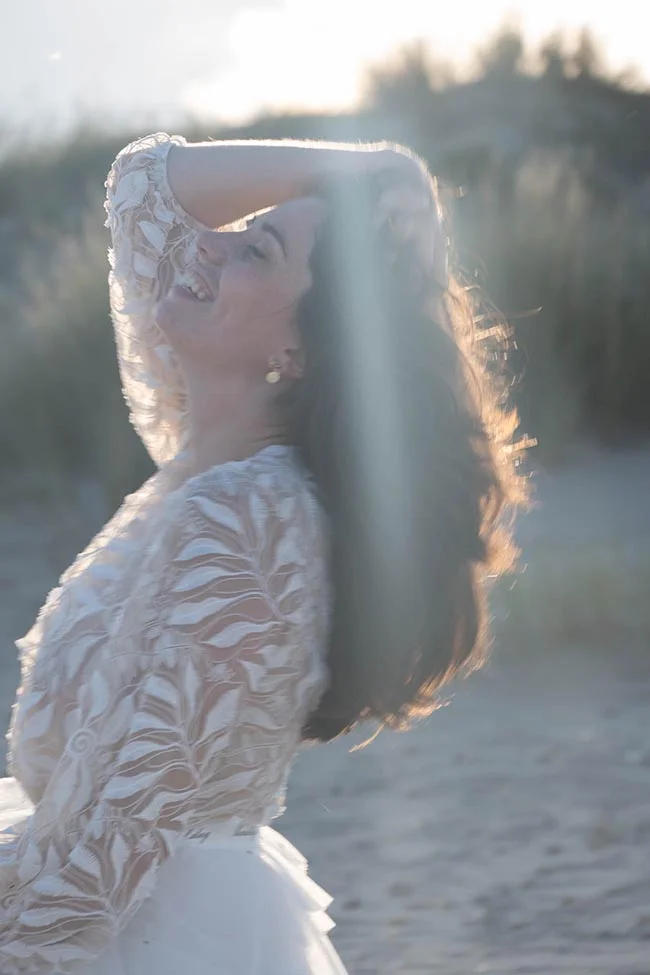 Mariée souriante en robe de dentelle blanche, cheveux au vent, baignée de lumière dorée sur une plage.