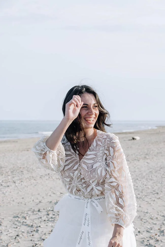 Femme souriante en tenue de mariée bohème blanche et dentelle, posant sur une plage lumineuse.