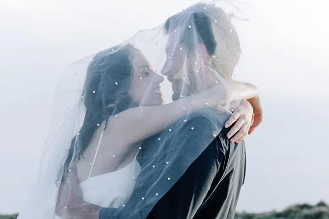 Couple d'amoureux enlacés sous un voile blanc translucide, ambiance romantique et lumineuse sur la plage.