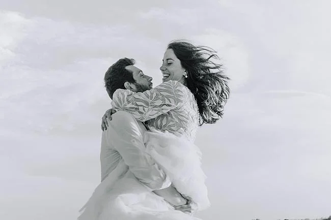 Couple de mariés s'enlaçant avec joie sur une plage, portrait en noir et blanc à l'ambiance romantique.