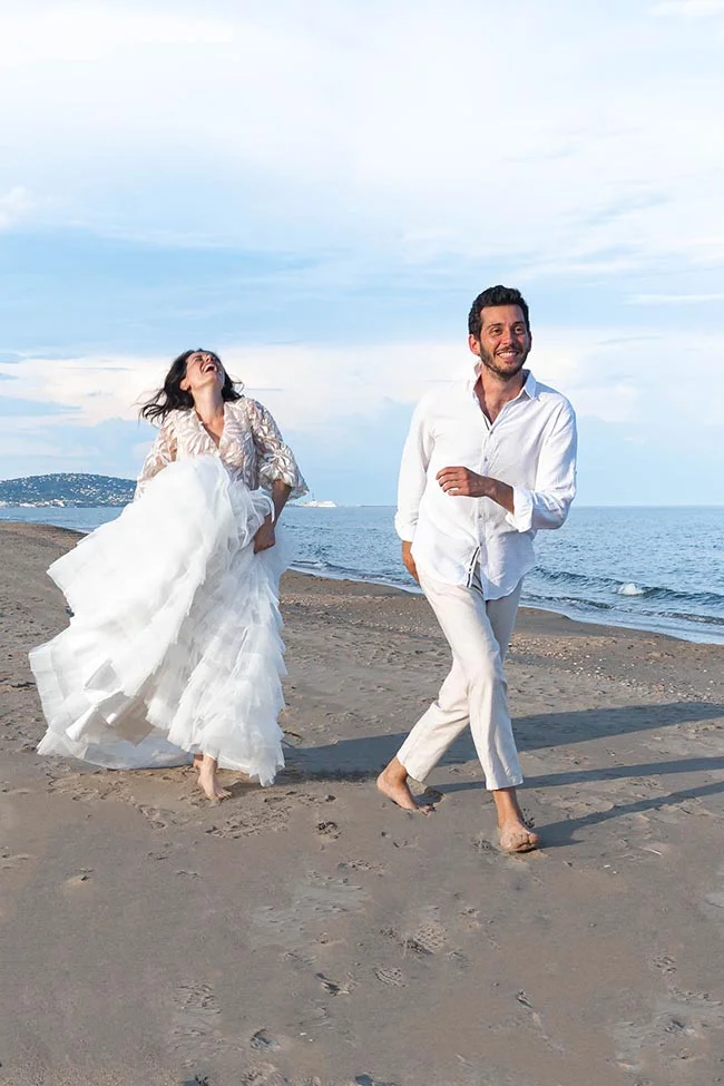 Un couple joyeux en tenues blanches court pieds nus sur une plage de sable fin sous un ciel clair.