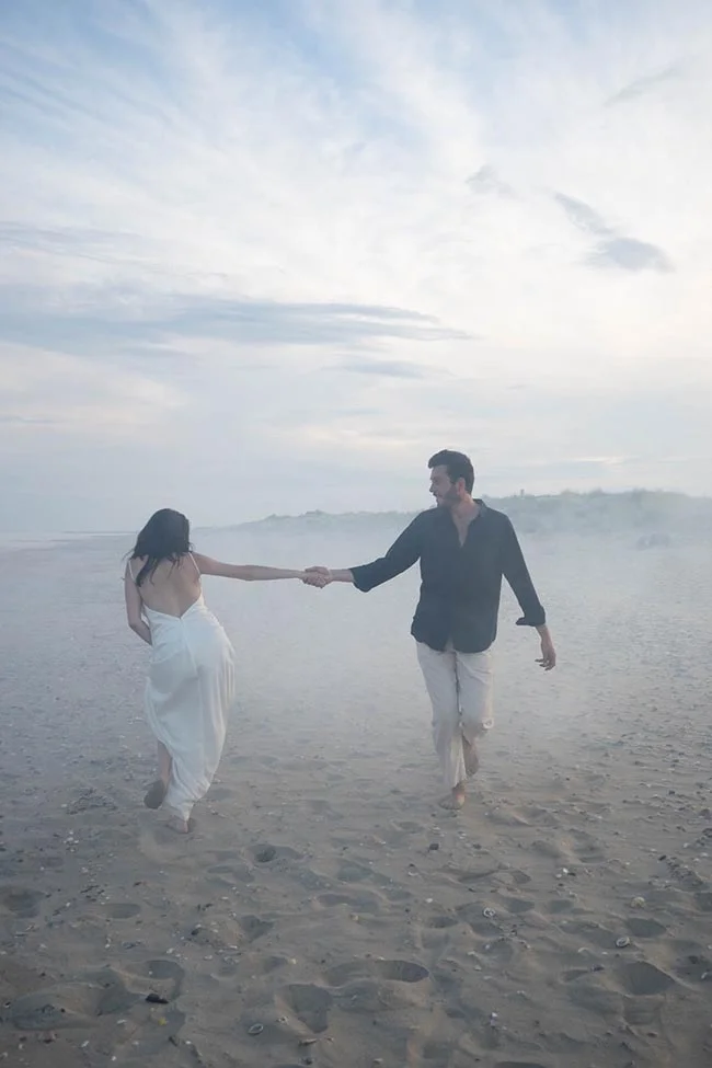 Un couple main dans la main marche sur une plage brumeuse, vêtus de blanc et de noir sous un ciel pâle.