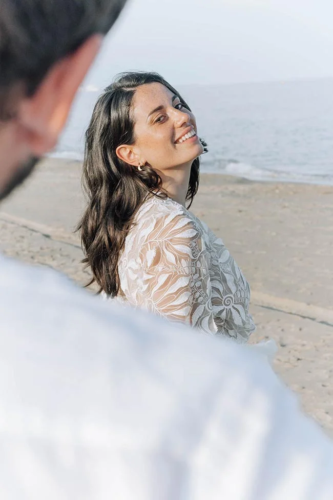 Une mariée souriante en robe blanche dentelée regarde son partenaire sur une plage lumineuse au bord de la mer.