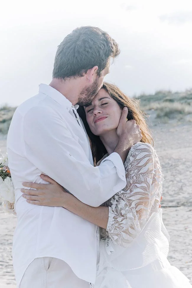 Couple en tenue blanche s'enlaçant tendrement sur une plage lumineuse dans une ambiance romantique.
