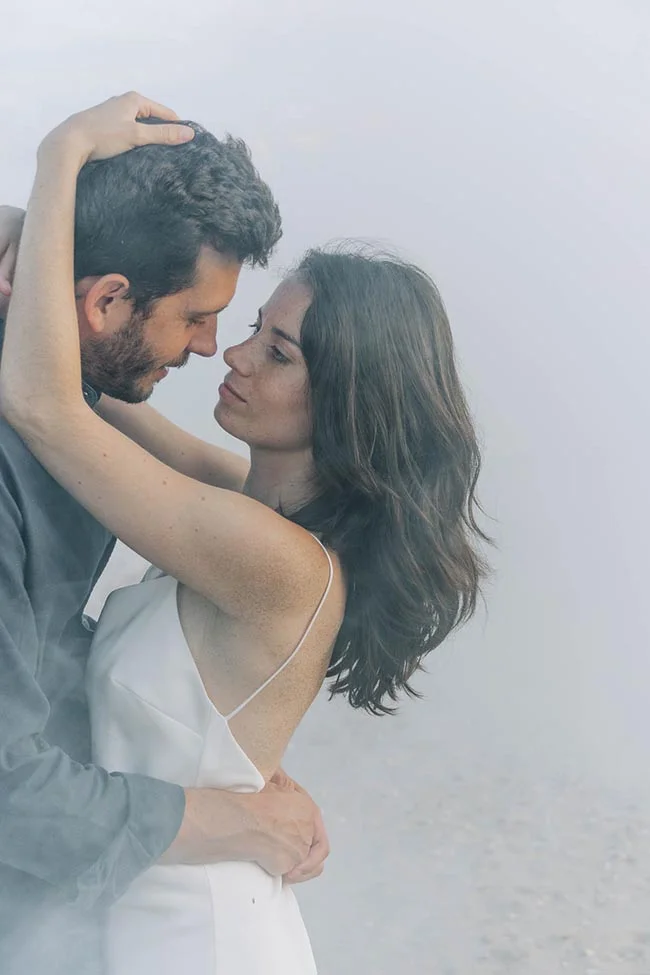 Couple en tenue de mariage s'enlaçant tendrement dans un décor brumeux et minimaliste aux tons clairs.