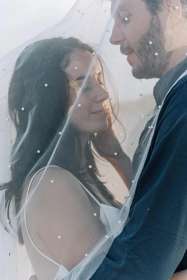 Couple s'enlaçant tendrement sous un voile blanc perlé, dans une ambiance douce et lumineuse en bord de mer.