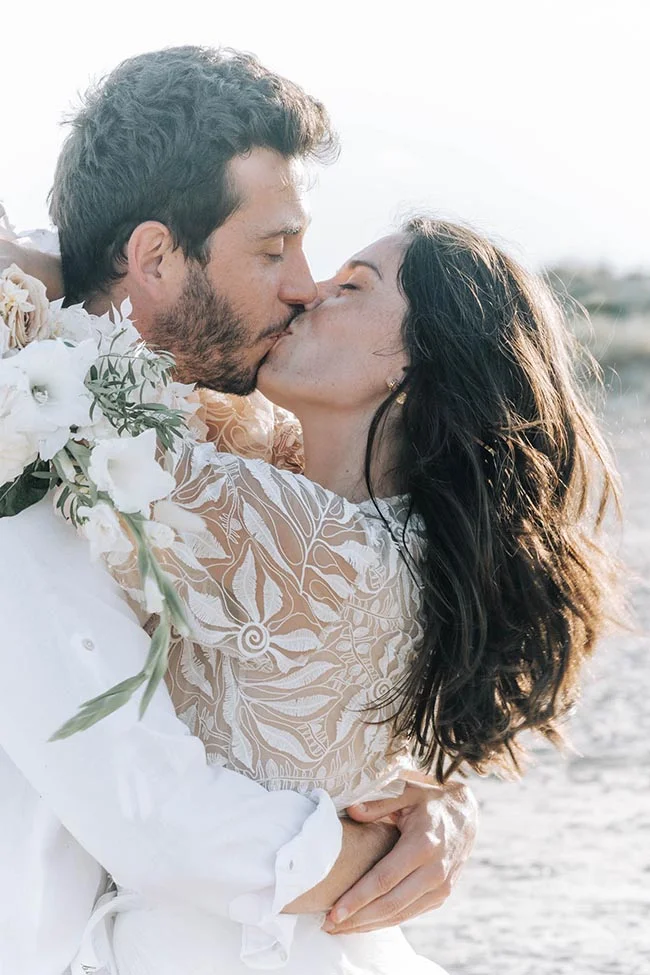 Un couple de mariés s'embrasse tendrement sur une plage, vêtus de blanc dans une ambiance lumineuse et romantique.