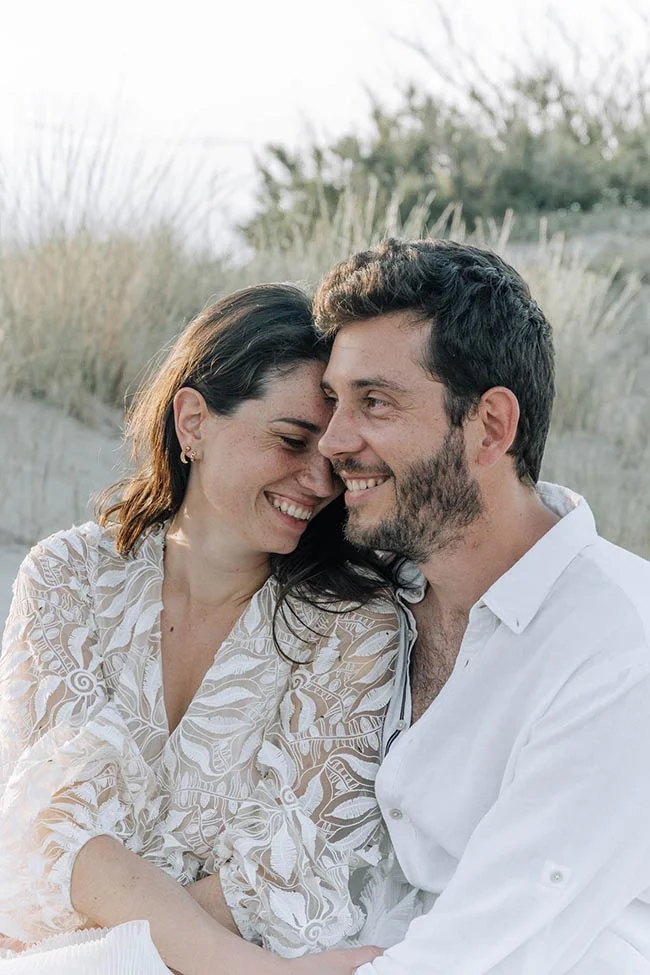 Couple souriant et complice en tenues blanches, s'enlaçant tendrement sur une plage aux tons clairs et naturels.
