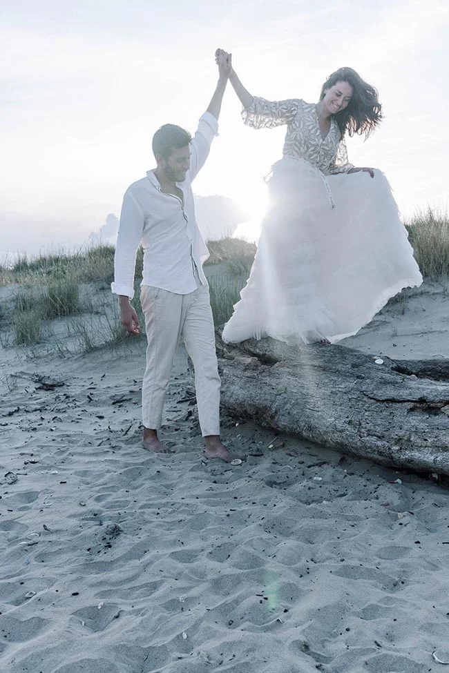 Couple marié s'enlaçant main dans la main sur une plage sablonneuse au coucher du soleil, ambiance romantique et lumineuse.