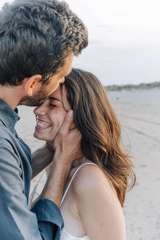 Couple complice s'enlaçant tendrement sur une plage au coucher du soleil dans des tons doux et naturels.