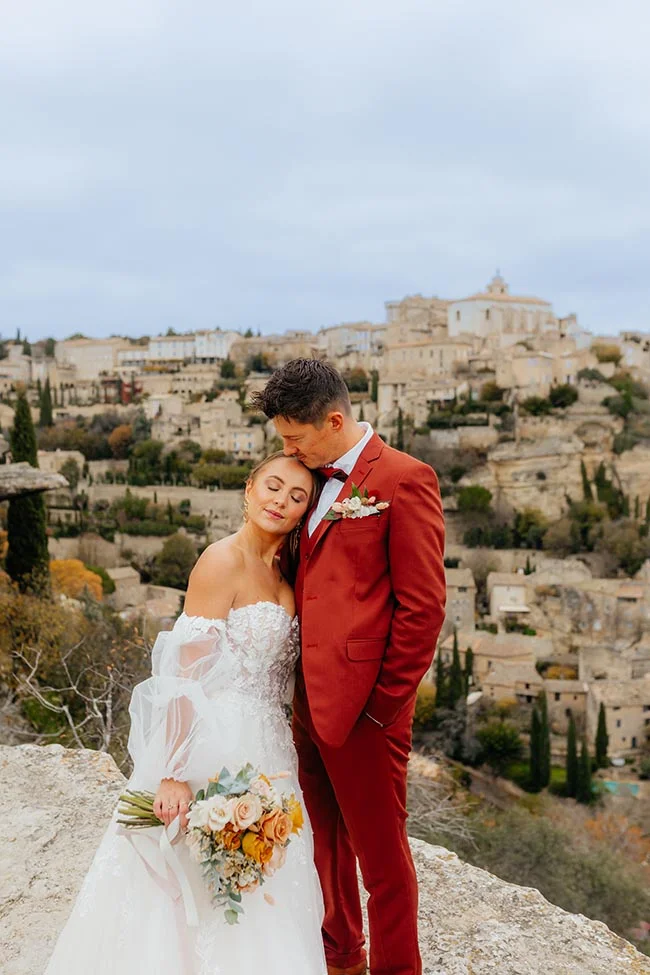 Jeunes mariés s'enlaçant tendrement devant un village provençal, elle en robe blanche et lui en costume rouge terracotta.
