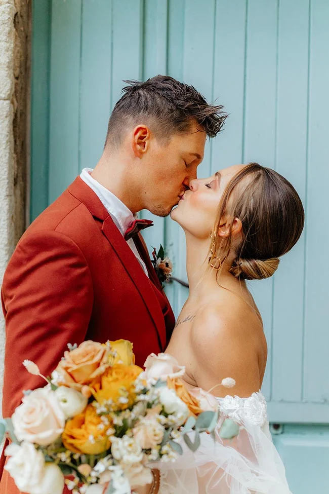 Jeunes mariés s'embrassant devant une porte bleue, le marié en costume terracotta et la mariée avec un bouquet coloré.