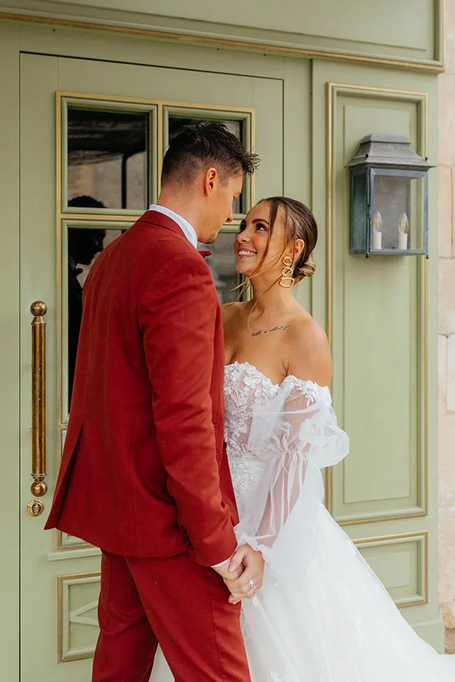 Jeunes mariés complices devant une porte pastel, lui en costume terracotta et elle en robe blanche vaporeuse.