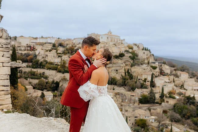 Jeunes mariés s'embrassant devant un village provençal, lui en costume rouge et elle en robe blanche.