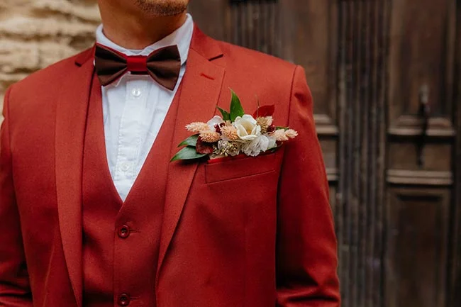Gros plan d'un marié portant un costume rouge brique avec nœud papillon et une boutonnière fleurie.