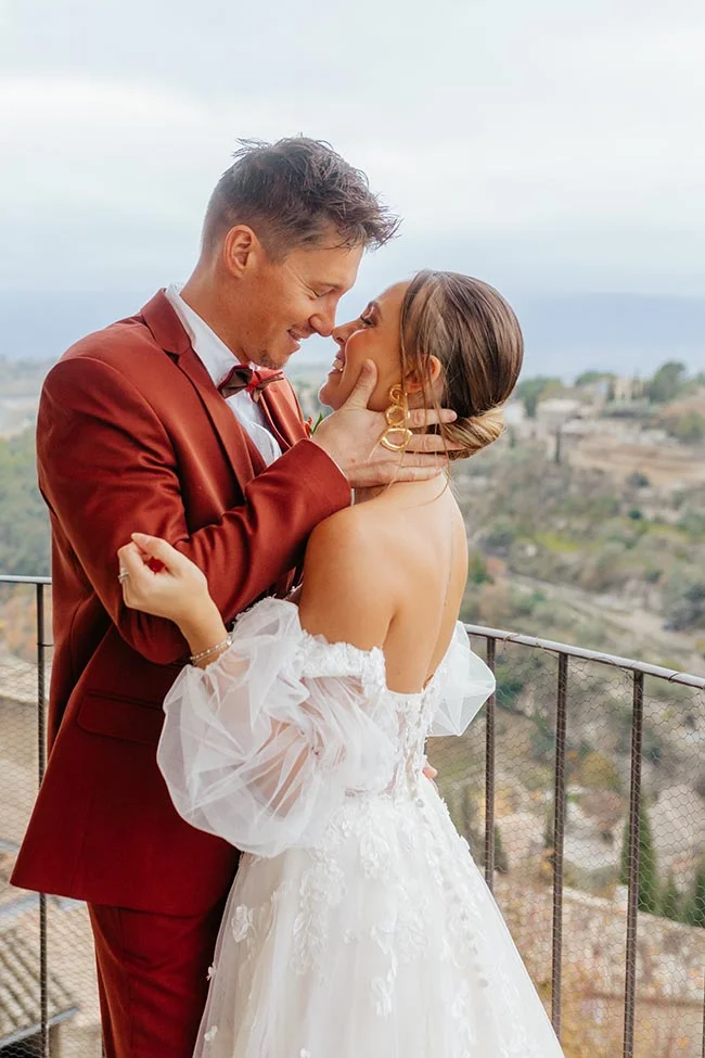 Couple complice en tenue de mariage, costume rouge et robe blanche, s'enlaçant face à un paysage provençal.