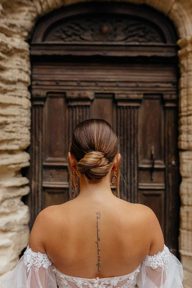 Dos d'une mariée en robe blanche avec un tatouage, face à une porte ancienne en bois dans un village provençal.