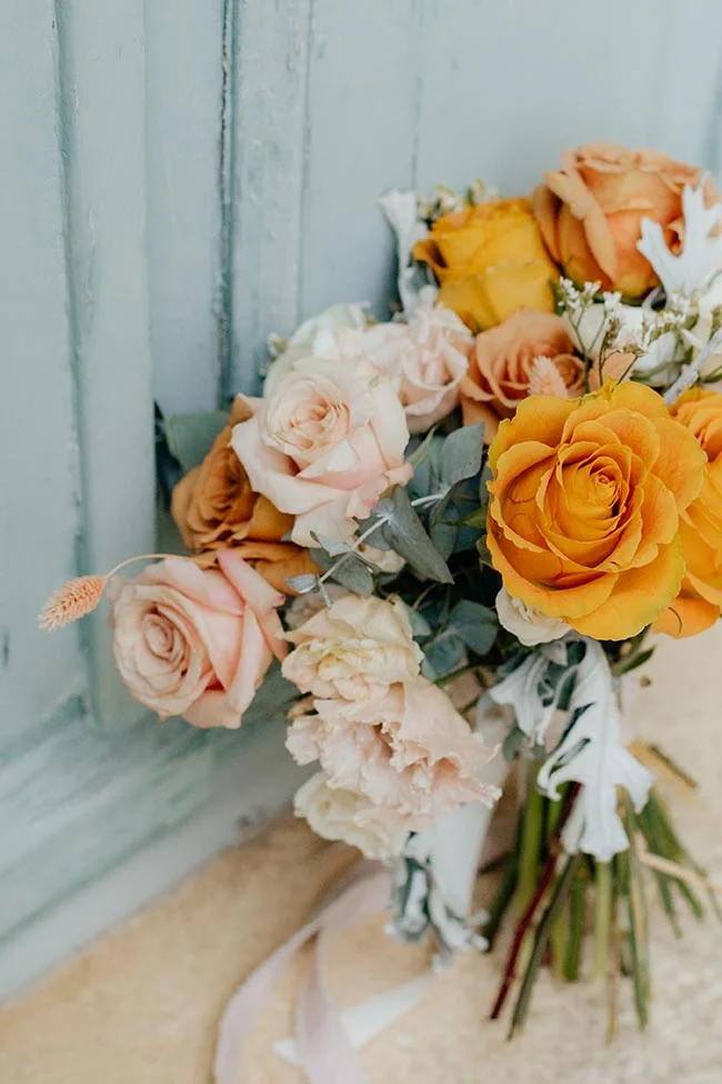 Bouquet de roses aux tons pêche, crème et ocre posé contre un mur bleu pastel, ambiance romantique et provençale.