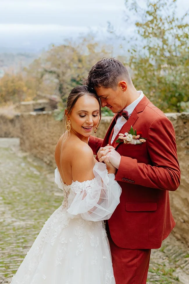 Jeunes mariés complices dans un village provençal, elle en robe blanche et lui en costume terracotta.