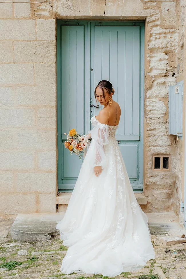 Mariée en robe blanche et bouquet coloré devant une porte bleue dans un village provençal.