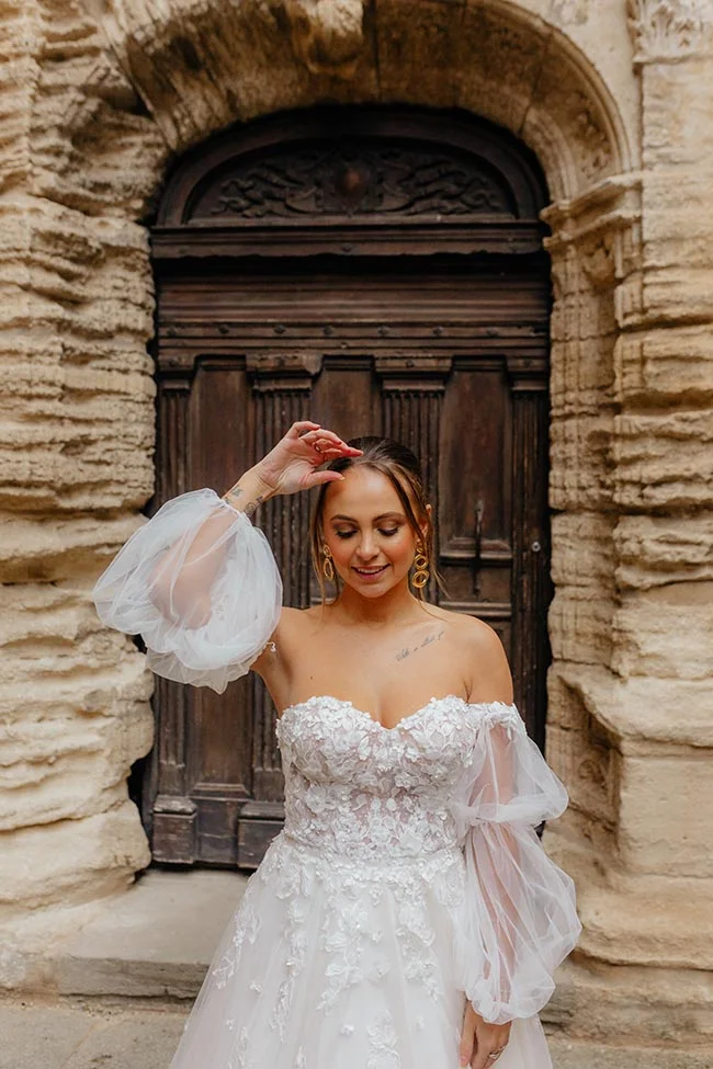 Mariée en robe blanche romantique avec manches bouffantes devant une porte ancienne en bois et pierre en Provence.
