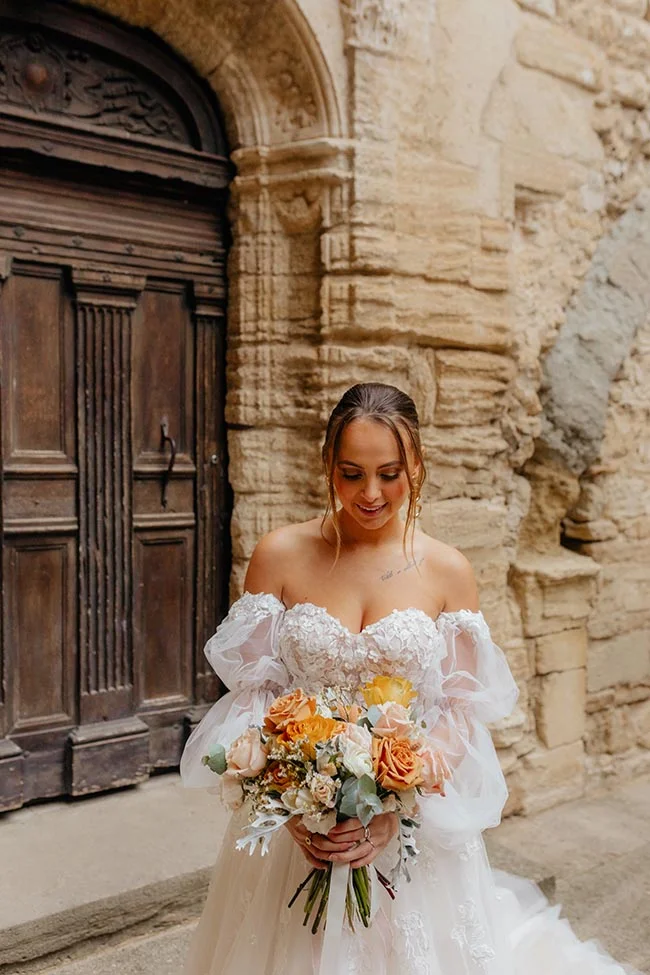 Mariée souriante en robe blanche et bouquet orangé devant une porte ancienne en bois et un mur de pierre provençal.