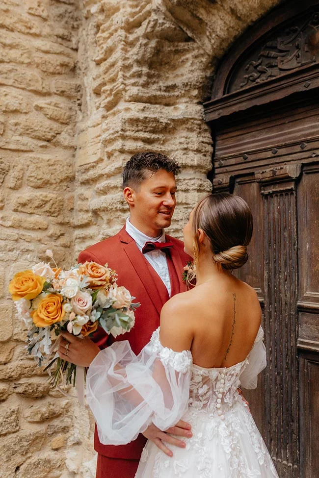 Couple marié s'enlaçant devant une porte ancienne, le marié en costume rouge et la mariée en robe blanche dentelée.