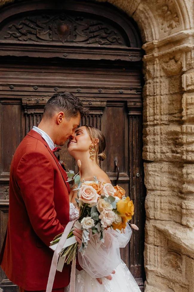 Couple marié s'embrassant tendrement devant une porte ancienne, avec un bouquet pastel et un costume rouge brique.