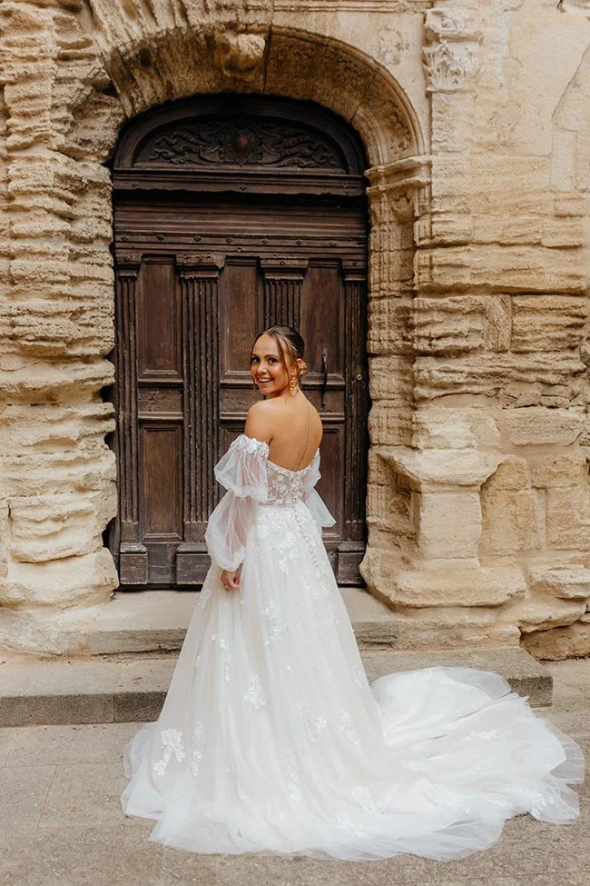 Une mariée en robe blanche vaporeuse pose devant une porte en bois sculpté et un mur en pierre ancienne.