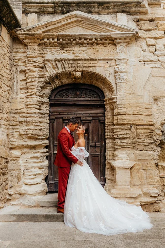 Couple de mariés s'embrassant devant une porte ancienne en pierre, costume rouge et robe blanche vaporeuse.