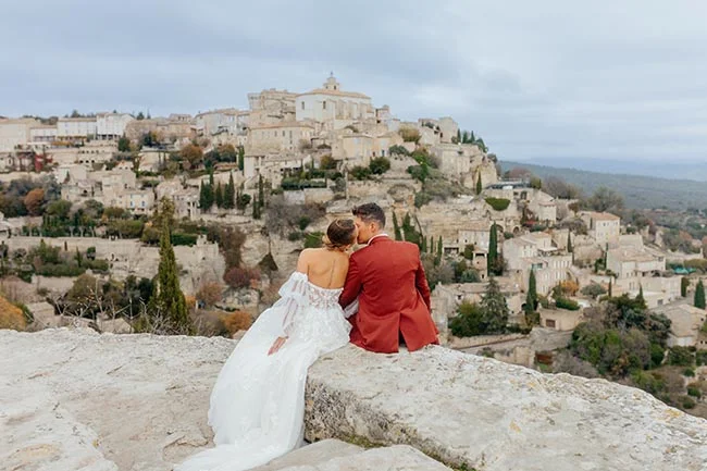Jeunes mariés s'embrassant face à un village provençal, elle en robe blanche et lui en costume rouge.