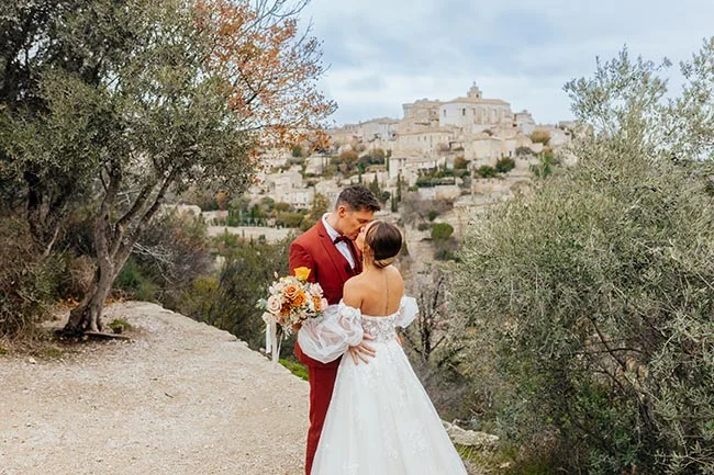 Jeunes mariés s'embrassant devant un village provençal, avec un costume rouge et une robe blanche vaporeuse.