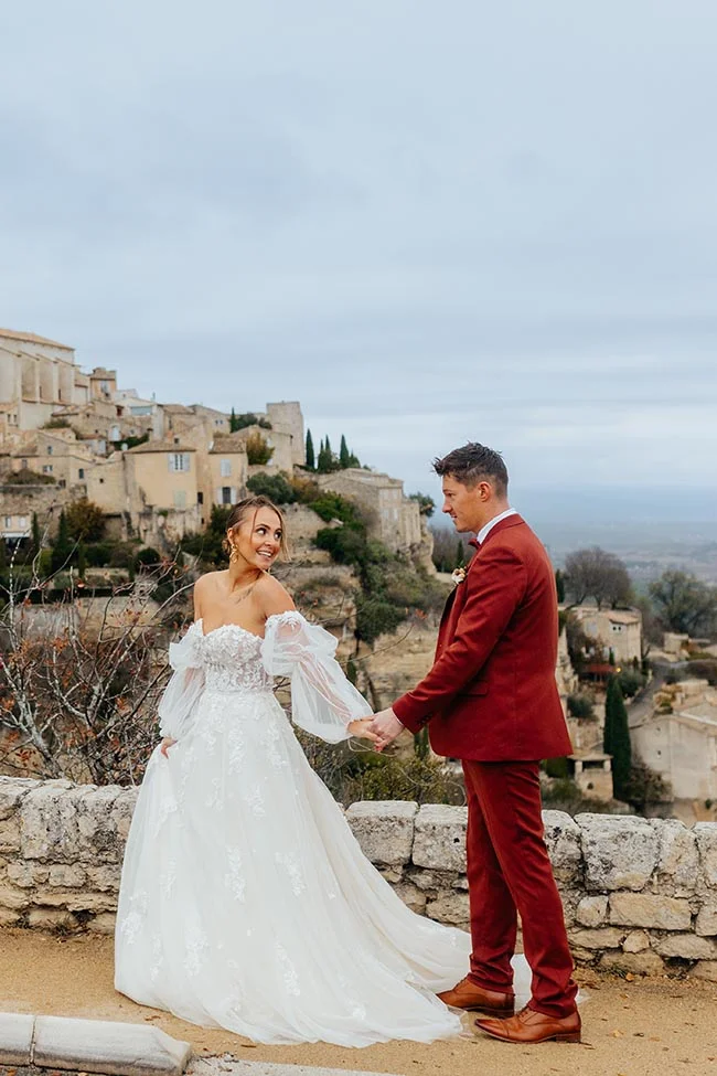 Couple de mariés main dans la main devant un village provençal, elle en robe blanche et lui en costume rouge brique.