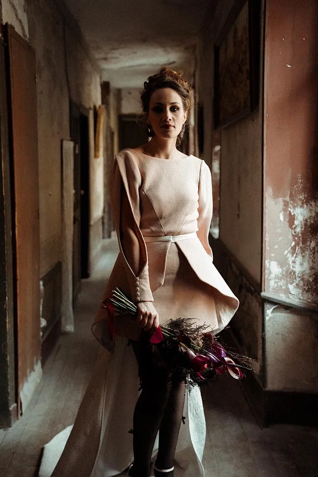 Femme en robe beige moderne tenant un bouquet sombre dans un couloir délabré, ambiance contrastée entre ombre et lumière.