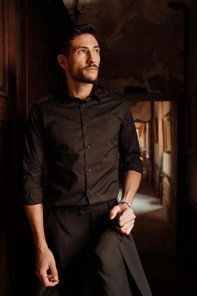 Homme élégant en chemise et pantalon noirs, posant dans un couloir sombre aux jeux d'ombre et de lumière.
