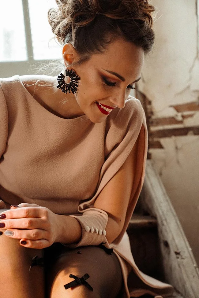 Femme souriante en robe beige et boucles d'oreilles noires, baignée d'une lumière douce et contrastée.
