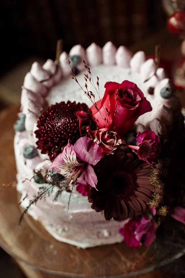 Gâteau de mariage blanc orné de fleurs bordeaux et roses, entouré de meringues, dans une ambiance sombre et romantique.