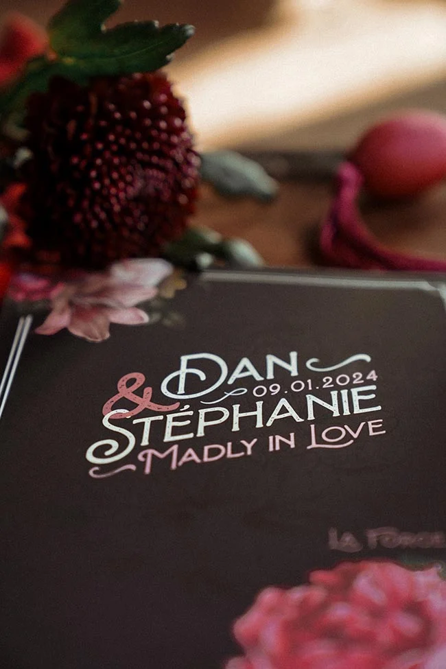 Gros plan sur un faire-part sombre et élégant pour Dan et Stéphanie, entouré de fleurs rouges dans une ambiance tamisée.