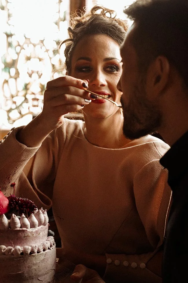 Une mariée souriante donne à goûter un gâteau à son époux dans une ambiance intime aux contrastes d'ombre et de lumière.