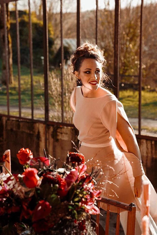 Femme élégante en robe beige posant dans une serre lumineuse, entourée de fleurs rouges aux tons chaleureux.