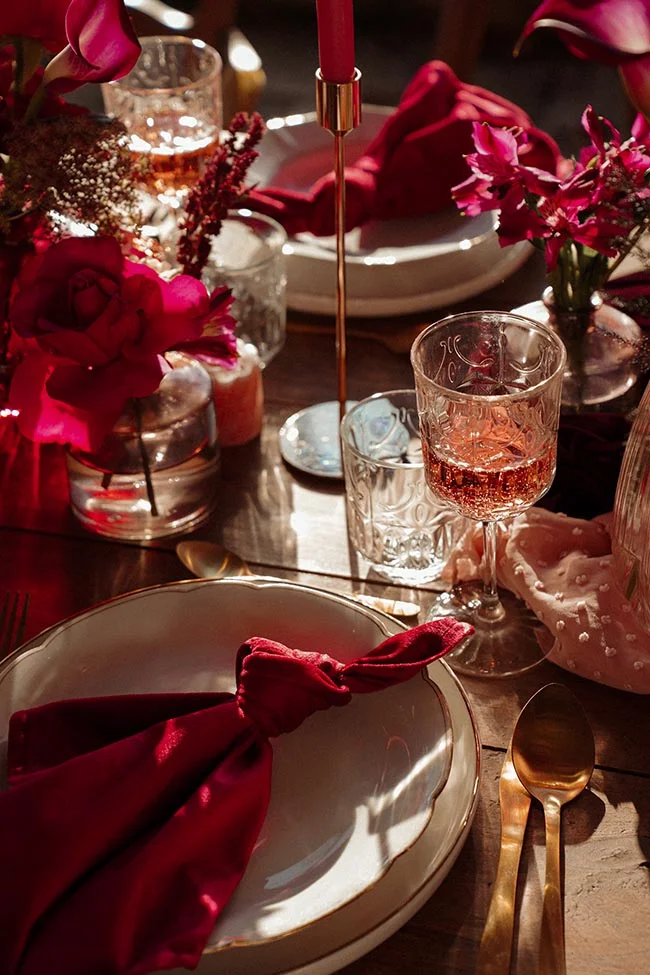 Table dressée avec des fleurs et serviettes rouge profond, verres ciselés et touches dorées sous une lumière contrastée.