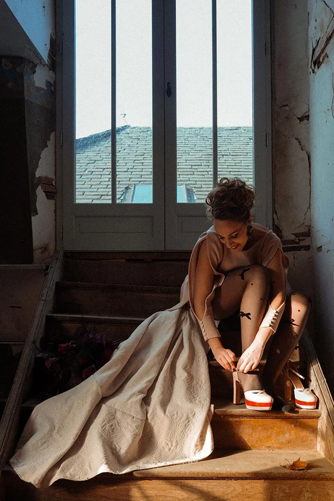 Une mariée en robe beige ajuste ses chaussures sur un escalier rustique, baignée d'une lumière naturelle contrastée.
