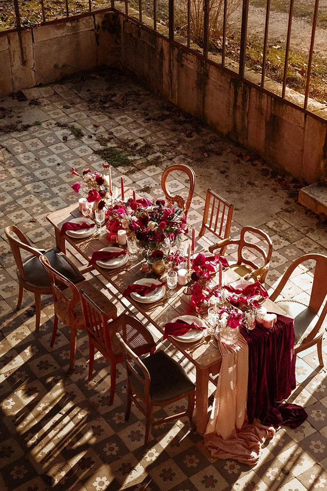 Table élégante dressée en extérieur avec fleurs bordeaux, bougies et chaises vintage sous un jeu d'ombre et de lumière.