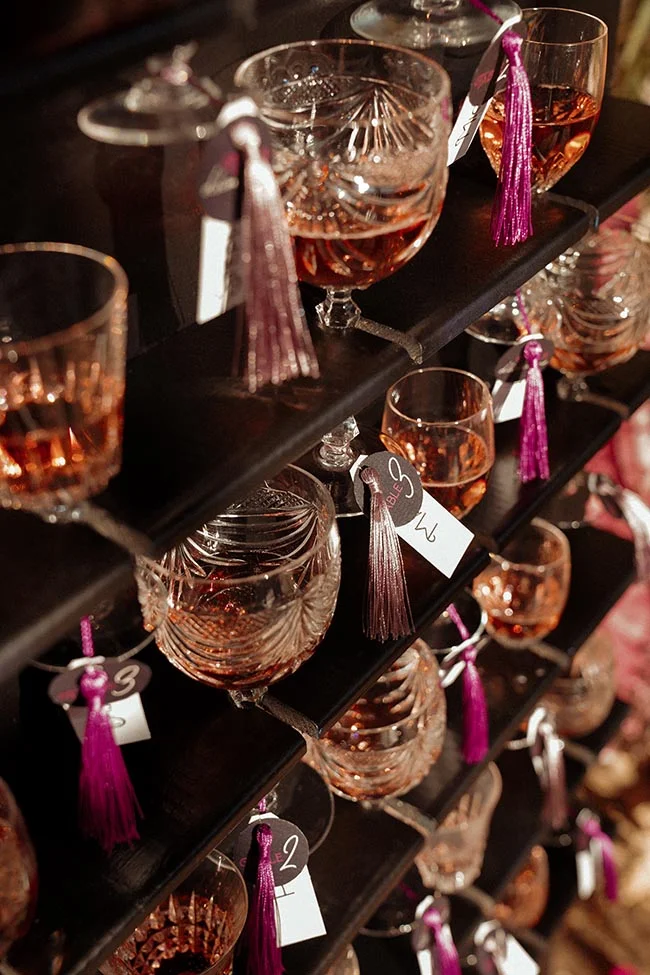 Verres à pied remplis de vin rosé, ornés de pompons mauves et d'étiquettes, disposés sur des étagères sombres.