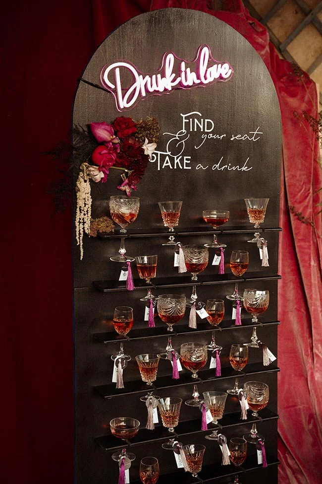 Mur de placement sombre avec néon rose, fleurs rouges et verres de cocktail pour un mariage élégant et mystérieux.