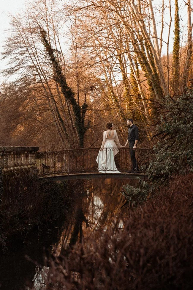 Couple de mariés se tenant la main sur un pont, entourés d'arbres aux tons automnaux dans une ambiance romantique.