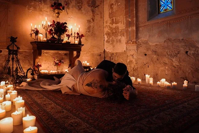 Couple s'embrassant au sol dans une pièce rustique éclairée à la bougie, ambiance romantique et mystérieuse.