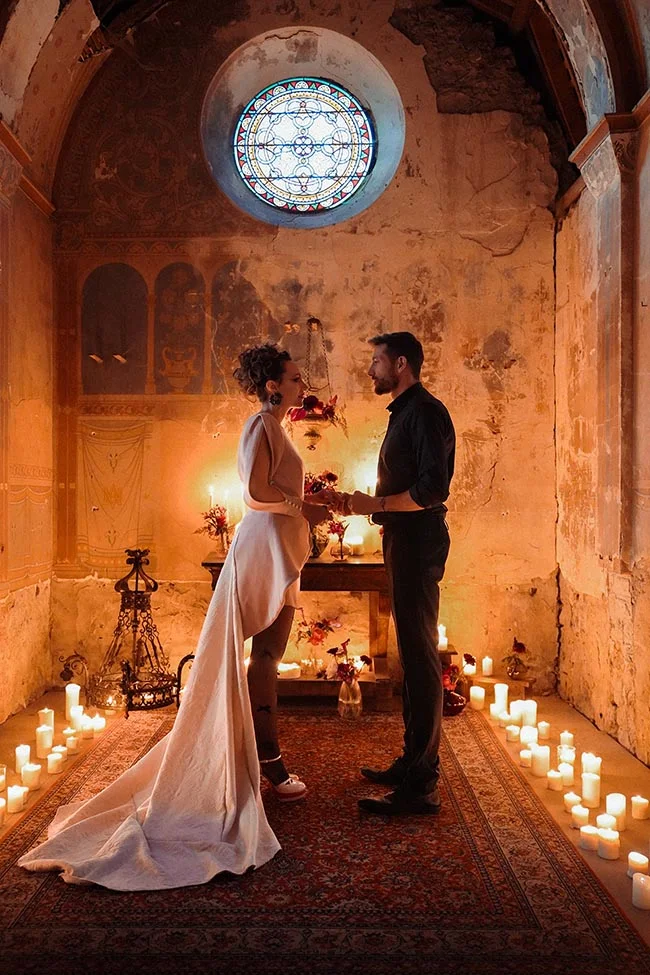 Couple en tenue de mariage se tenant la main dans une chapelle ancienne aux tons chauds et éclairée aux bougies.