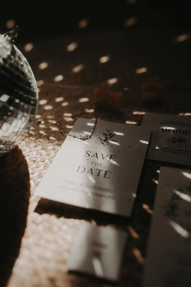 Faire-part de mariage minimaliste et boule à facettes sur un fond texturé, baignés d'une lumière solaire tamisée.